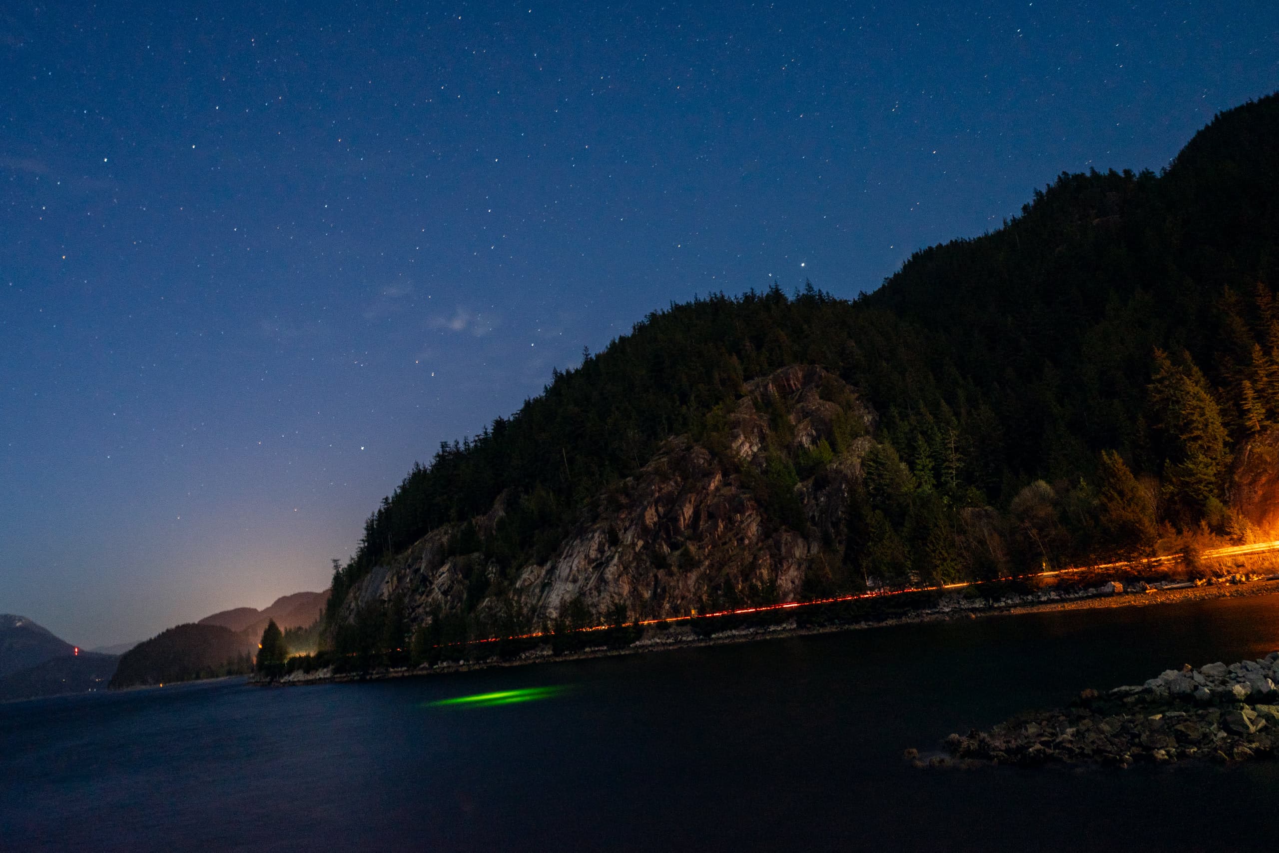 Starry night with mountains, water, and a light streak from cars