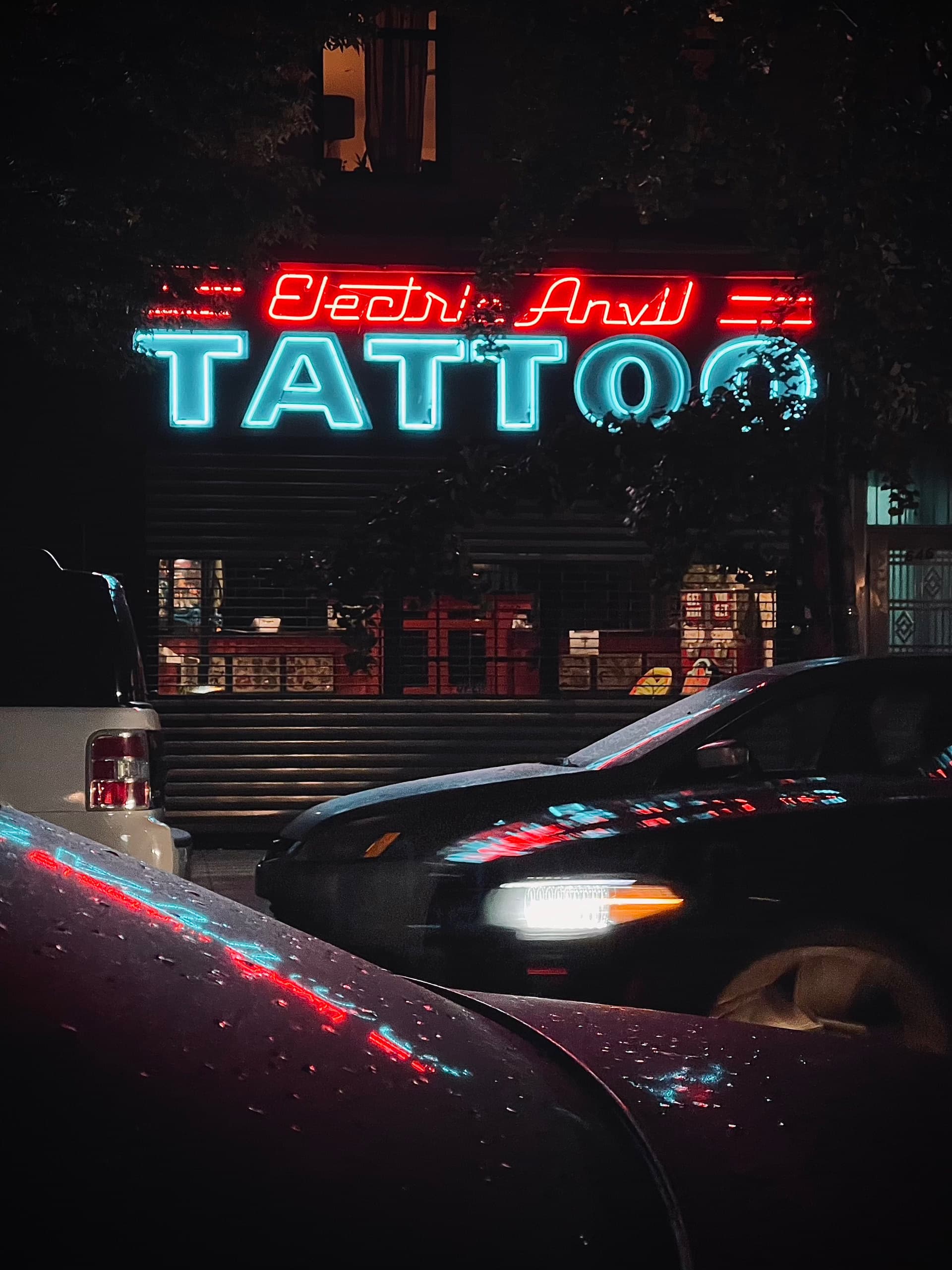 Red and cyan neon sign that says Electric Anvil Tattoo with reflection off two cars passing through