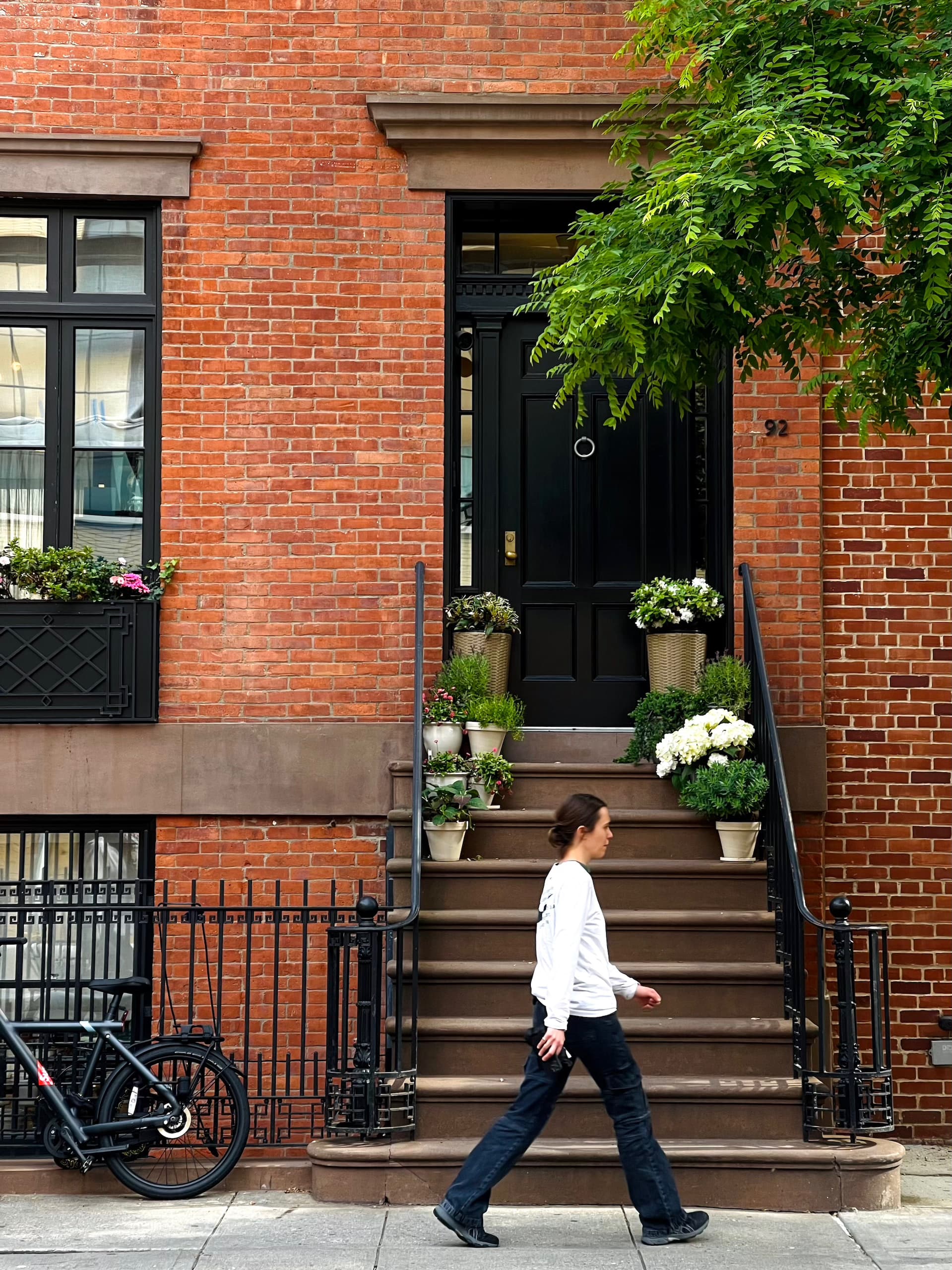 A woman in white shirt and jean walking right in front of an apartment with a brick wall on the street