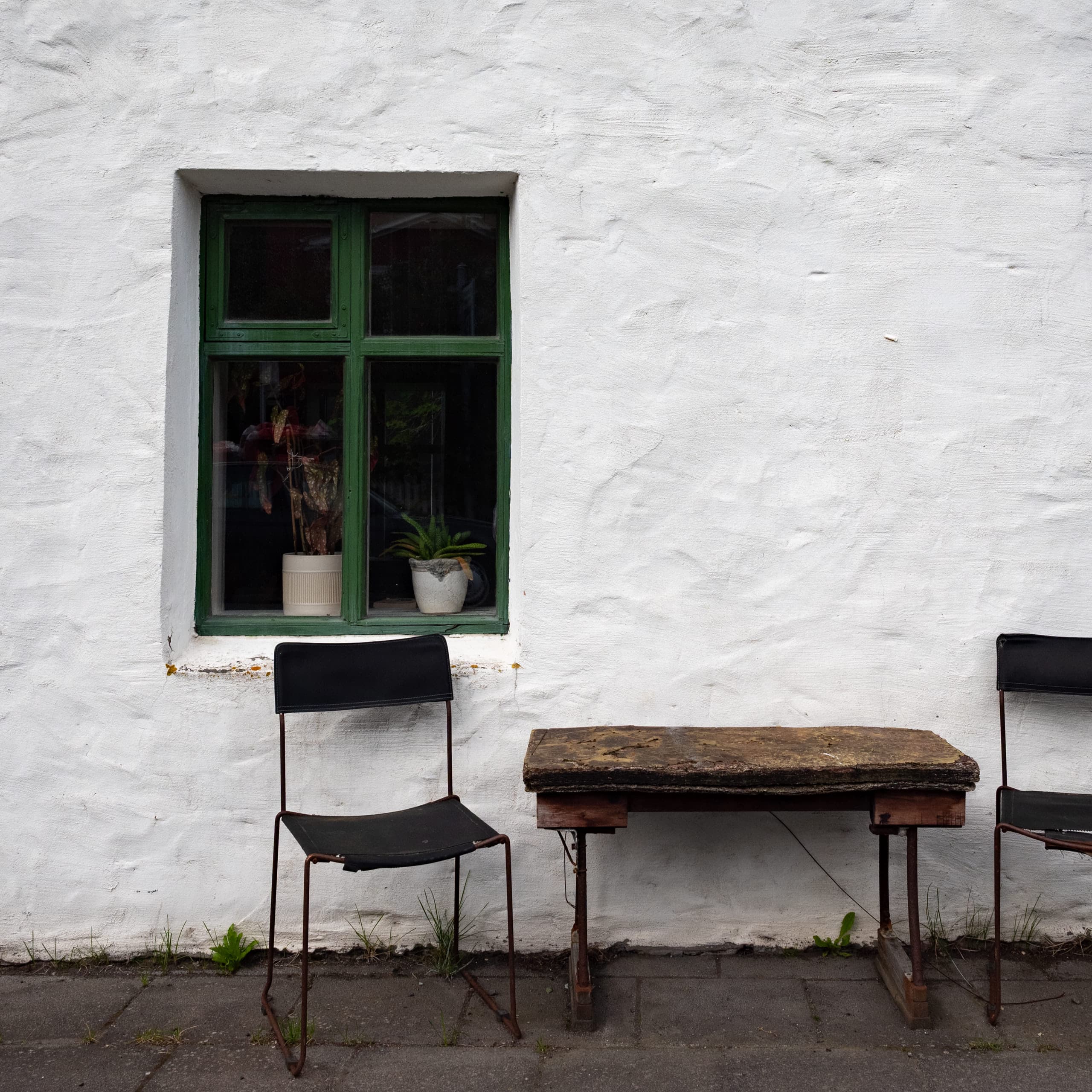 Two chairs and a table in front of a white wall and a window on the left with two plants