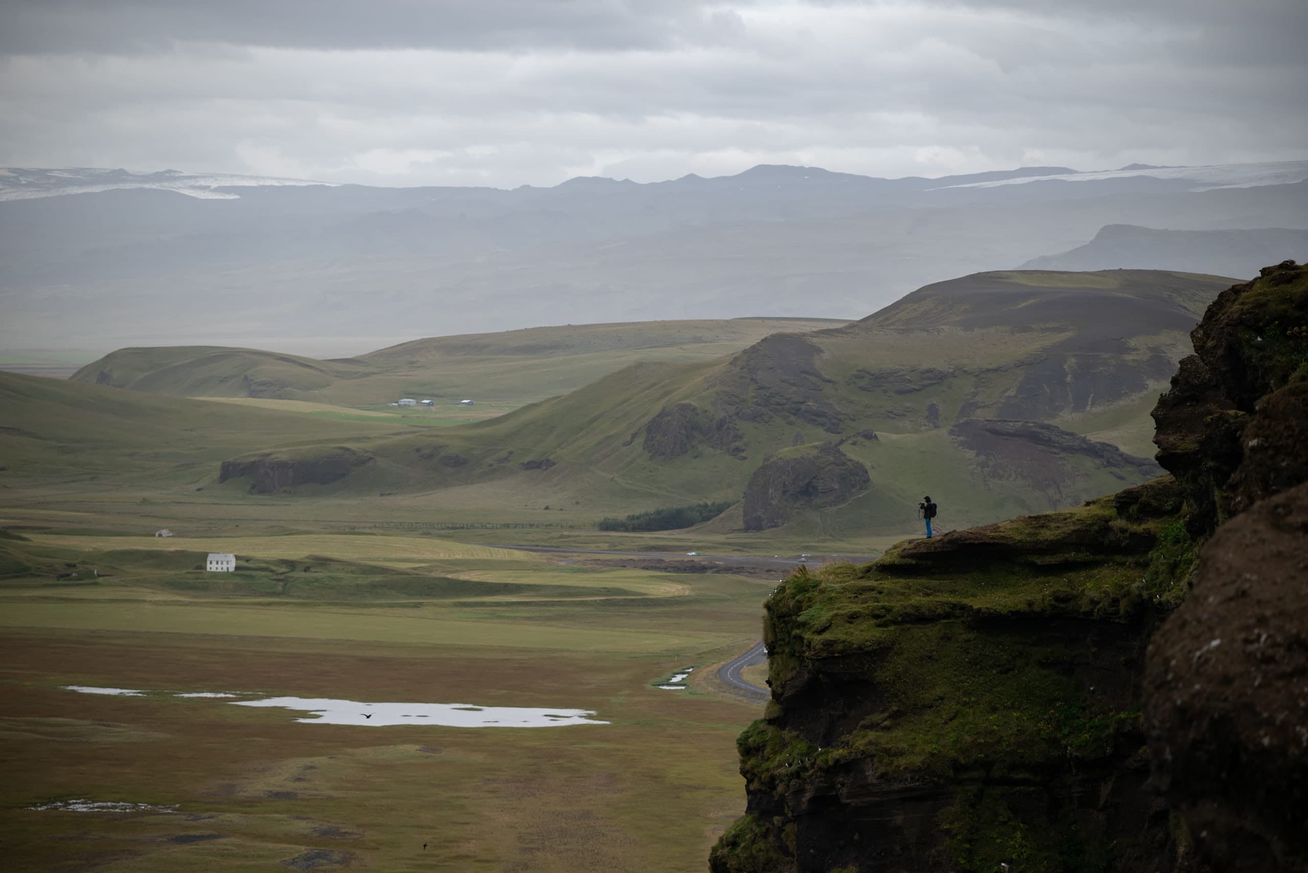 A photographer standing on a cliff on the bottom-right overlooking a vast landscape of Iceland in the background