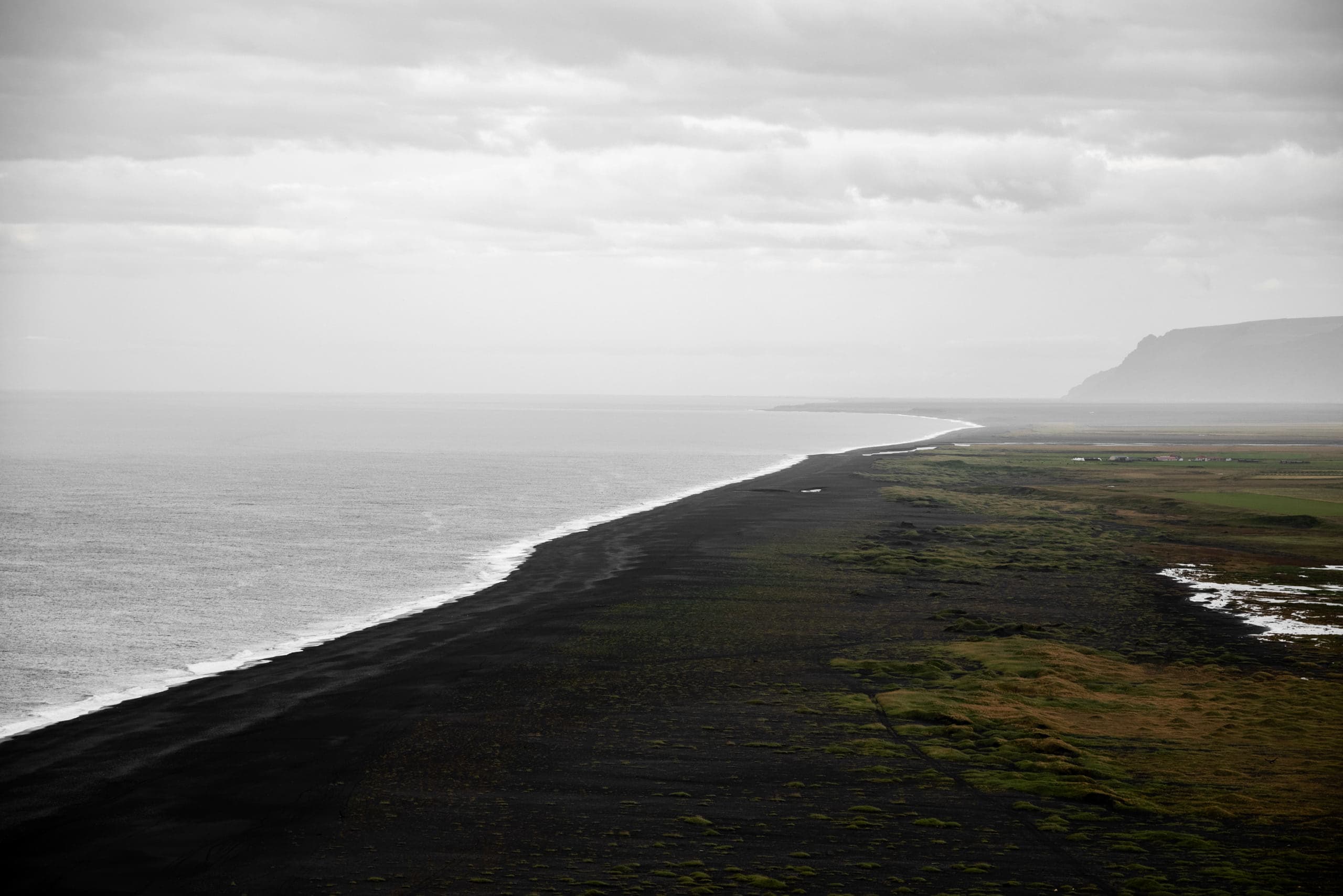 A landscape with the ocean on the left and a black sand beach on the right stretching toward the top-right of the frame