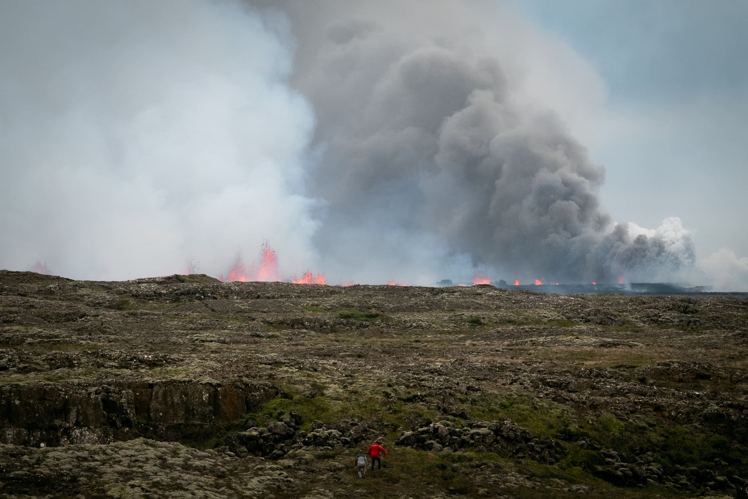 2 people walking toward a fissure eruption at a distance, with huge plumes of smoke filling in the background