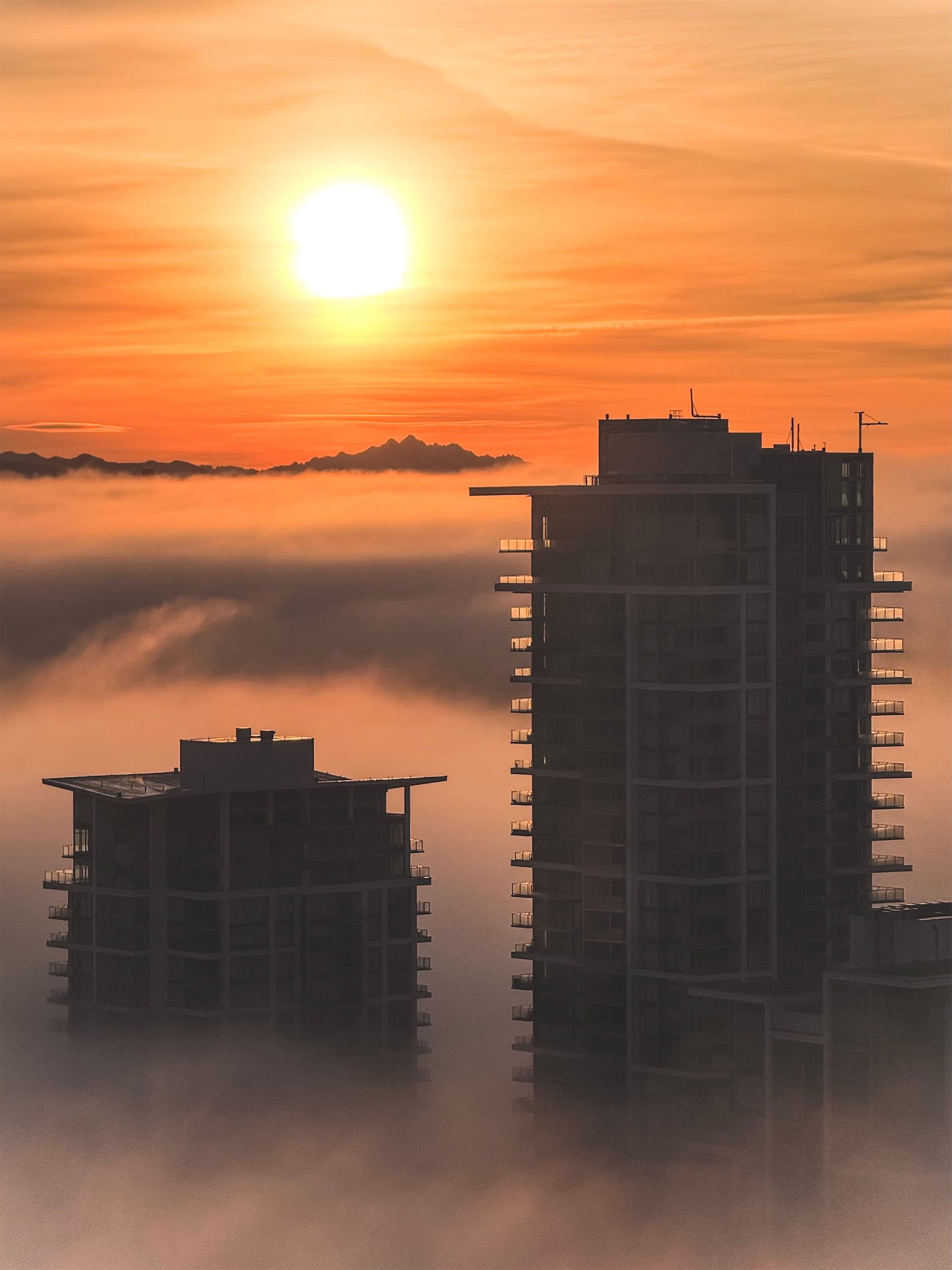 Two condo towers in a sea of clouds, a visible mountain range in the background, and a sun rising above the horizon